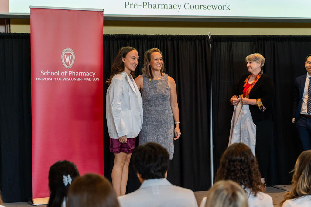 Photo by Paulius Musteikis A student poses with Kate Rotzenberg in front of a red School of Pharmacy banner while Karen Kopacek holds a coat awaiting the next student