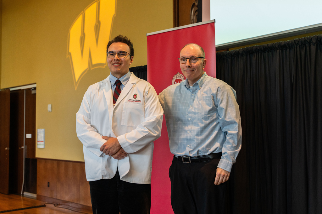 Photo by Paulius Musteikis A student poses in front of a red School of Pharmacy banner with an older man
