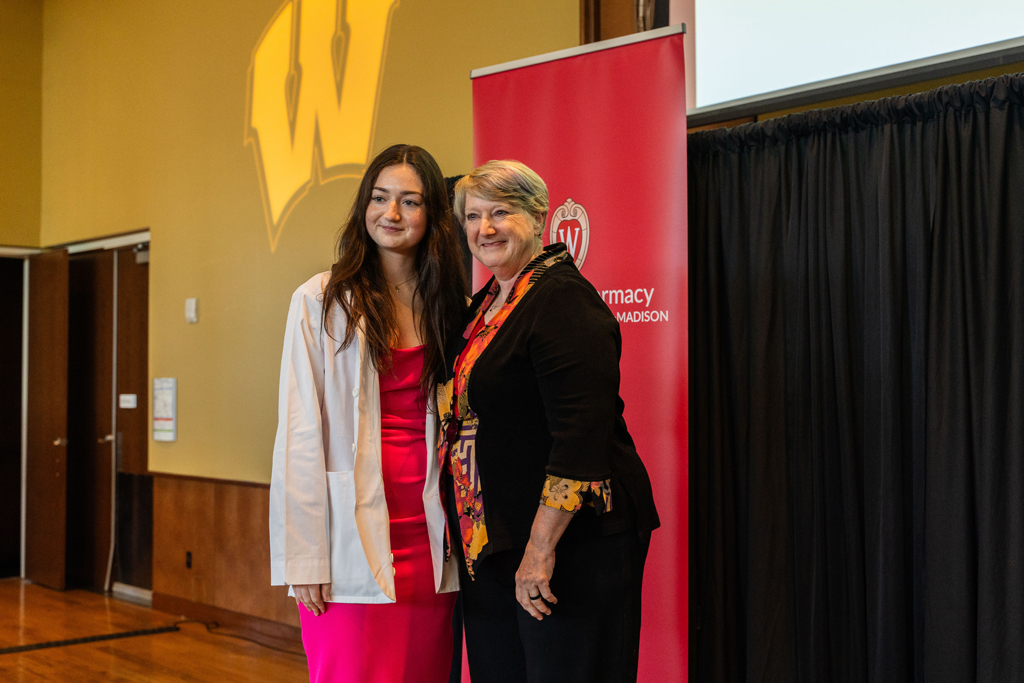 Photo by Paulius Musteikis A student in a white coat poses with Karen Kopacek in front of a red School of Pharmacy banner