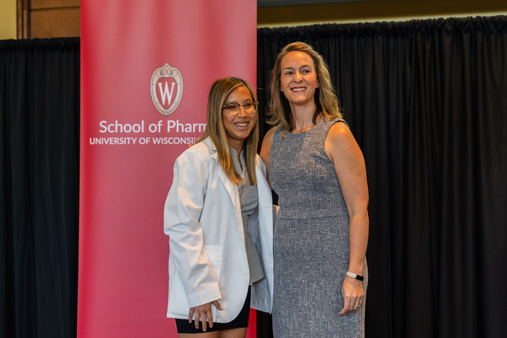 2024 White Coat Ceremony A student poses with Kate Rotzenberg in front of the red School of Pharmacy banner
