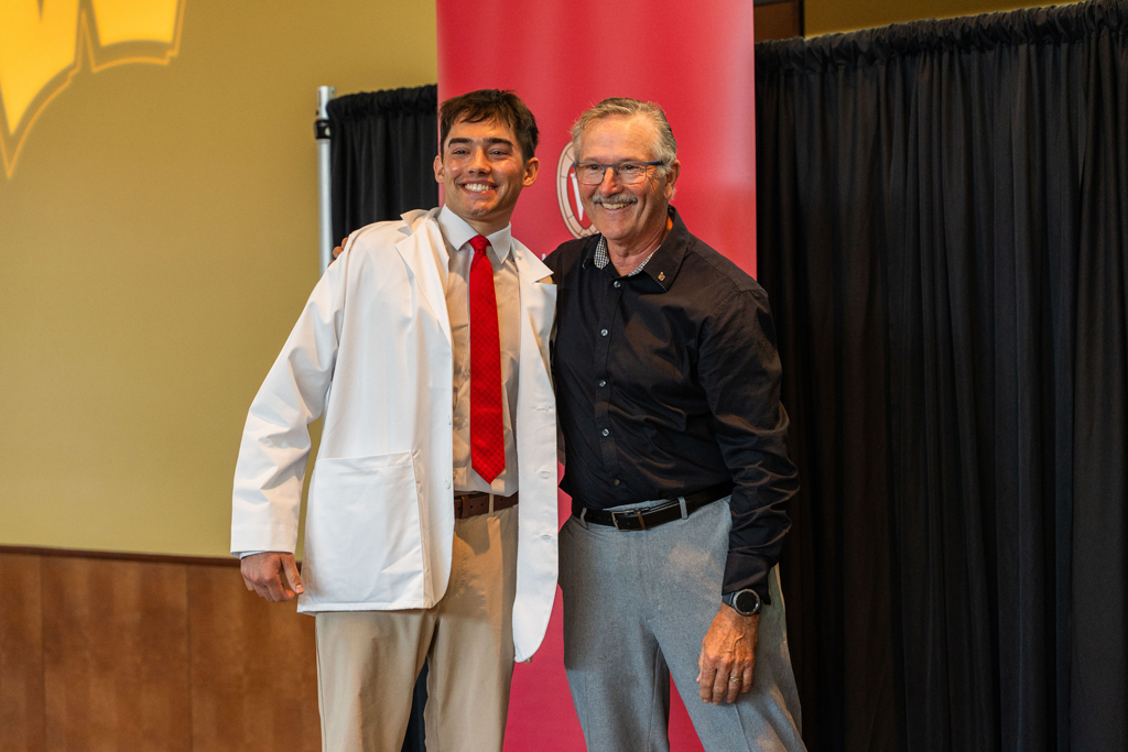 2024 White Coat Ceremony A student in a white coat and an older man smile and pose in front of a red School of Pharmacy banner