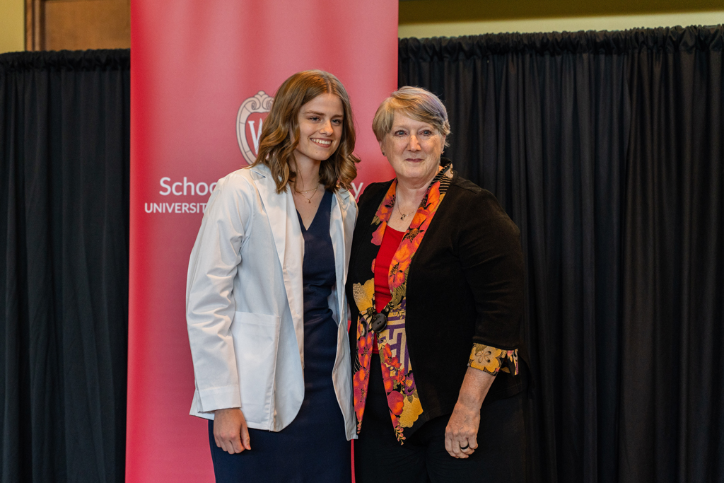Photo by Paulius Musteikis A student in a white coat poses with Karen Kopacek in front of a red School of Pharmacy banner