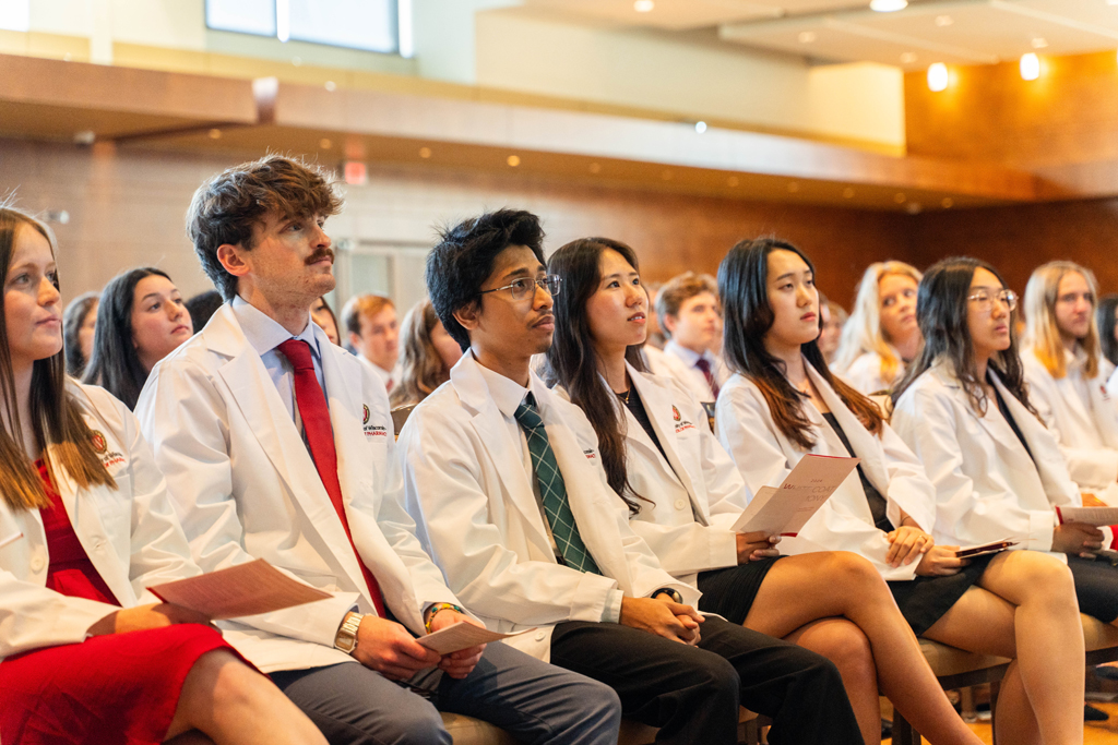 Photo by Paulius Musteikis Seated students in white coats listen in to a speaker