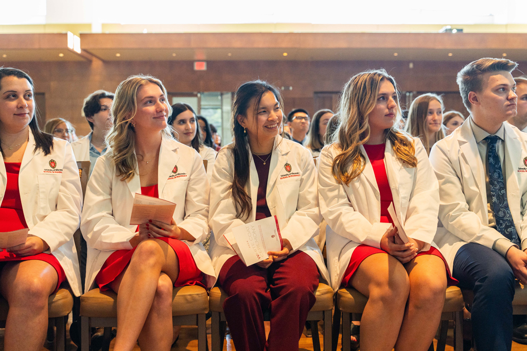 Photo by Paulius Musteikis Seated students in white coats smile and listen in to a speaker