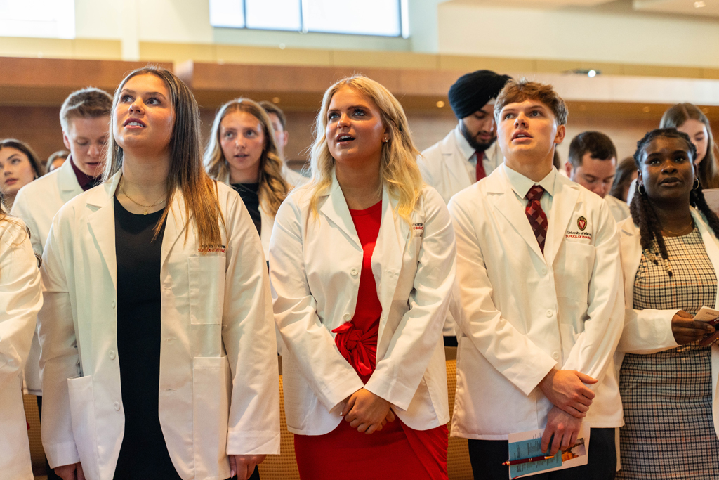 Photo by Paulius Musteikis Students in white coats read the Oath of a Pharmacist off of screens in front of the room