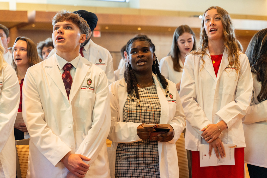 Photo by Paulius Musteikis Students in white coats read the Oath of a Pharmacist off of screens in front of the room