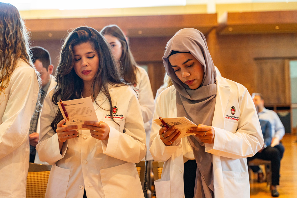 Photo by Paulius Musteikis Female students in white coats read the Oath of a Pharmacist off the back of their White Coat Ceremony program