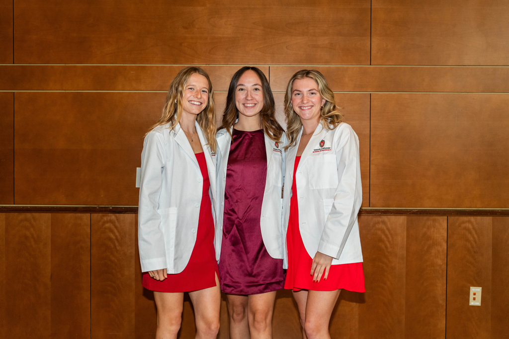 2024 White Coat Ceremony Three female students in white coats pose for a photo against a wooden wall