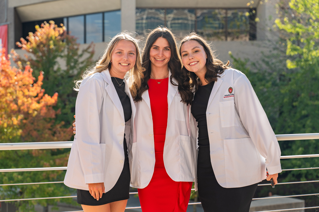 Photo by Paulius Musteikis Three female students in white coats smile for a photo outside