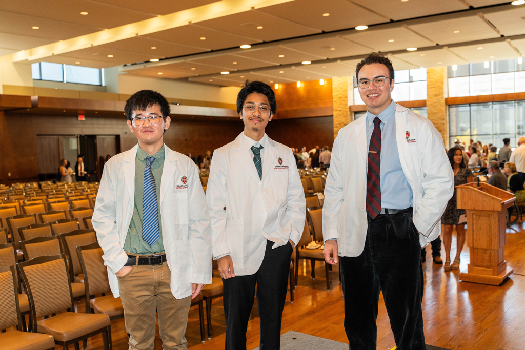 Photo by Paulius Musteikis Three male students in white coats pose in front of empty chairs
