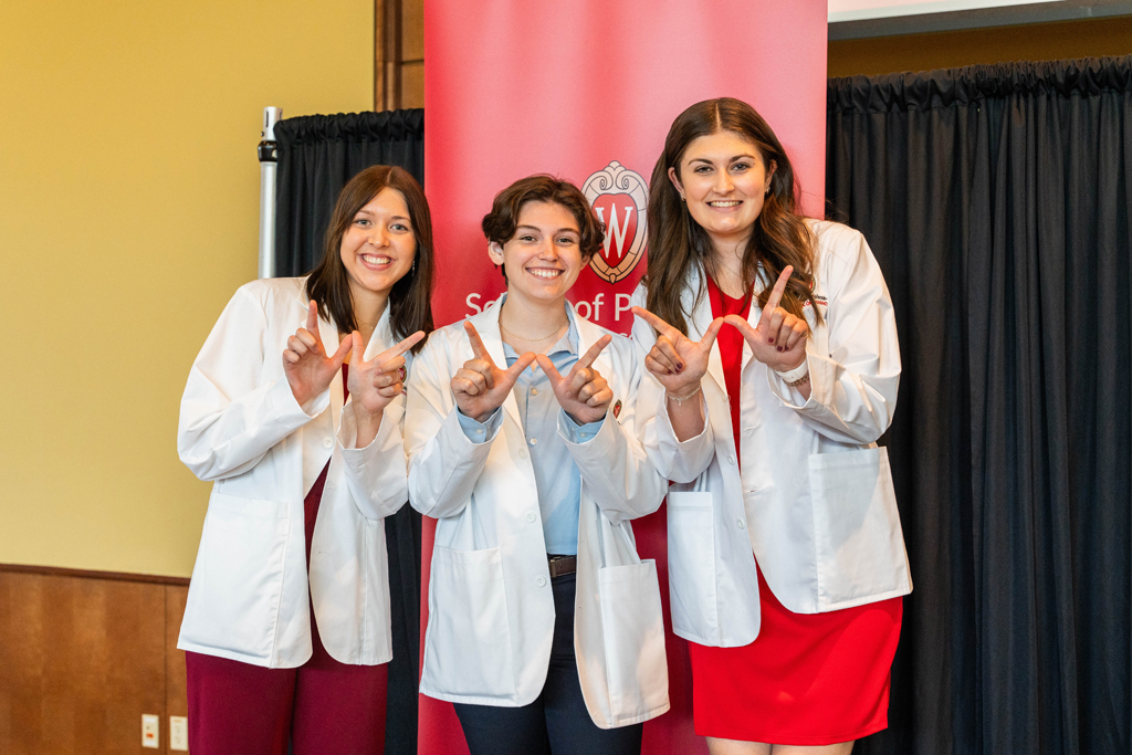 Photo by Paulius Musteikis Three students in white coats pose in front of red School of Pharmacy banner