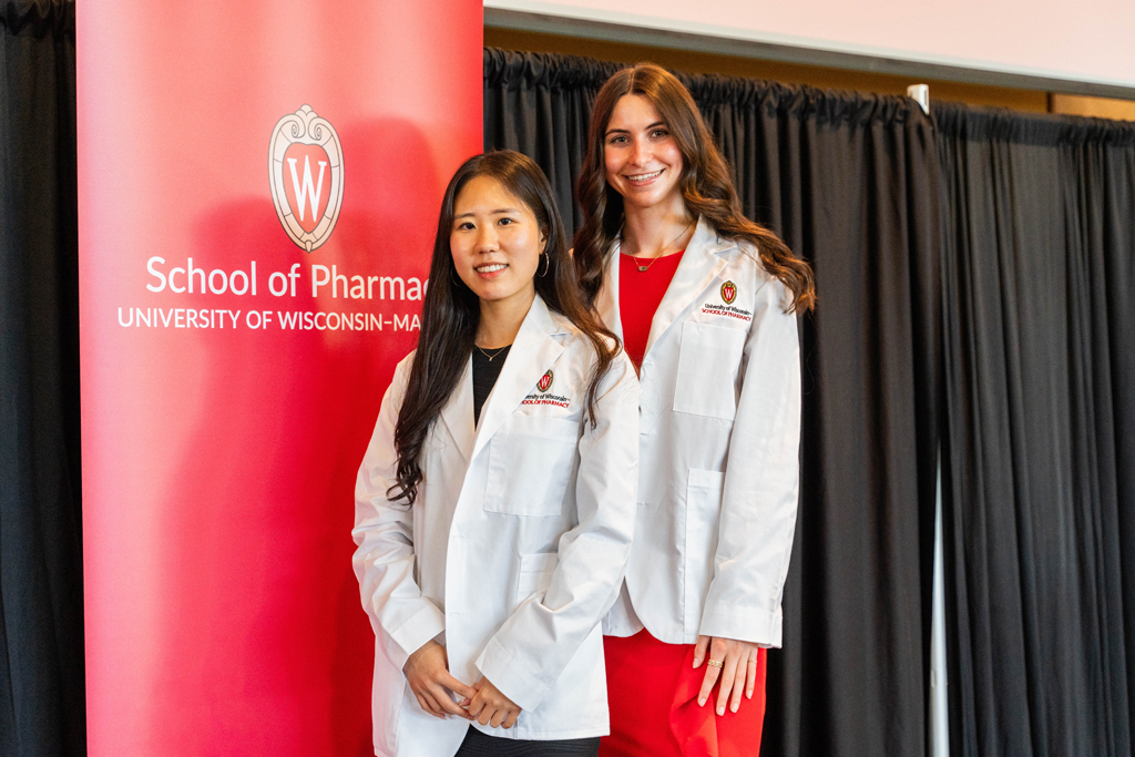 Photo by Paulius Musteikis Two female students in white coats pose in front of a red School of Pharmacy banner