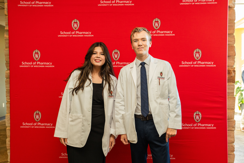 Photo by Paulius Musteikis A female and male student in white coats pose in front of red School of Pharmacy background