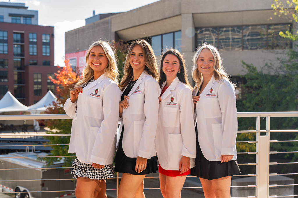 Photo by Paulius Musteikis Four female students pose outside and hold their white coat lapel to showcase the School of Pharmacy logo