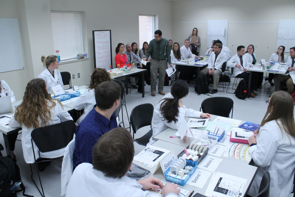 2019-PTx-Asthma-Lab-022 Instructor addressing room of students sitting at table filled with asthma supplies for lab
