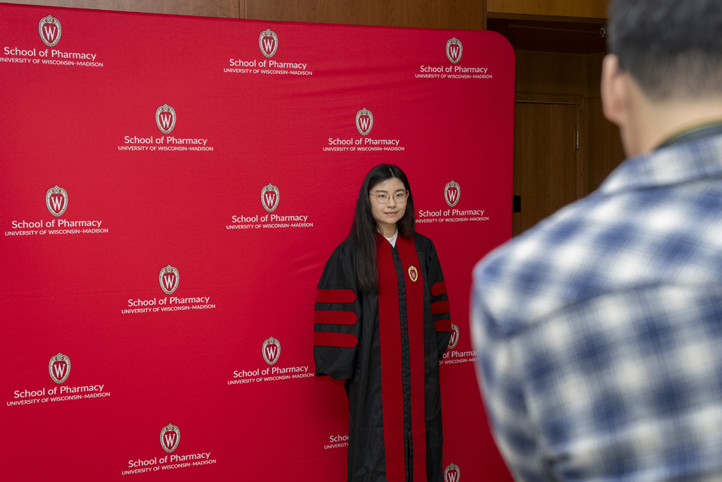 PharmD Graduation Awards Breakfast at the Pyle Center May 10, 2024. A graduate poses in front of a red School of Pharmacy background