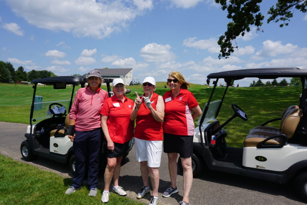 DSC07322 Four female alumni wearing red stand and smile in front of golf carts