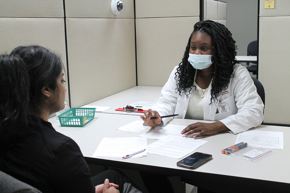 A student in a white coat speaks with a dark-haired person across a table.