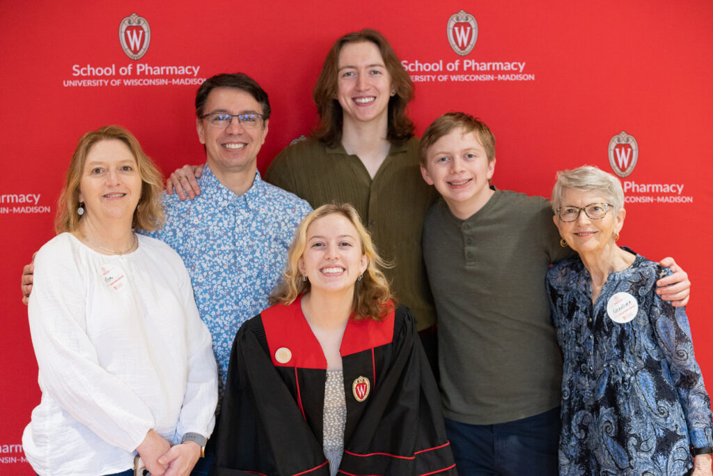 A PharmTox graduate and her family pose in front of the red School of Pharmacy backdrop.