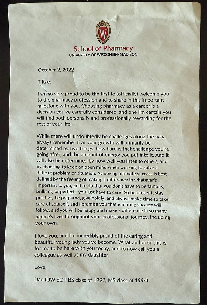 An image of a letter that reads as follows:
School of Pharmacy University of Wisconsin-Madison
October 2, 2022
T Rae:
I am so very proud to be the first to (officially) welcome you to the pharmacy profession and to share in this important milestone with you. Choosing pharmacy as a career is a decision you've carefully considered, and one I'm certain you will find both personally and professionally rewarding for the rest of your life.
While there will undoubtedly be challenges along the way, always remember that your growth will primarily be determined by two things: how hard is that challenge you're going after, and the amount of energy you put into it. And it will also be determined by how well you listen to others, and by choosing to keep an open mind when working to solve a difficult problem or situation. Achieving ultimate success is best defined by the feeling of making a difference in whatever's important to you, and to do that you don't have to be famous, brilliant, or perfect...you just have to care! So be present, stay positive, be prepared, give boldly, and always make time to take care of yourself, and I promise you that enduring success will follow, and you will be happy and make a difference in so many people's lives throughout your professional journey, including your own.
I love you, and I'm incredibly proud of the caring and beautiful young lady you've become. What an honor this is for me to be here with you today, and to now call you a colleague as well as my daughter.
Love,
Dad (UW SOP BS class of 1992, MS class of 1994)