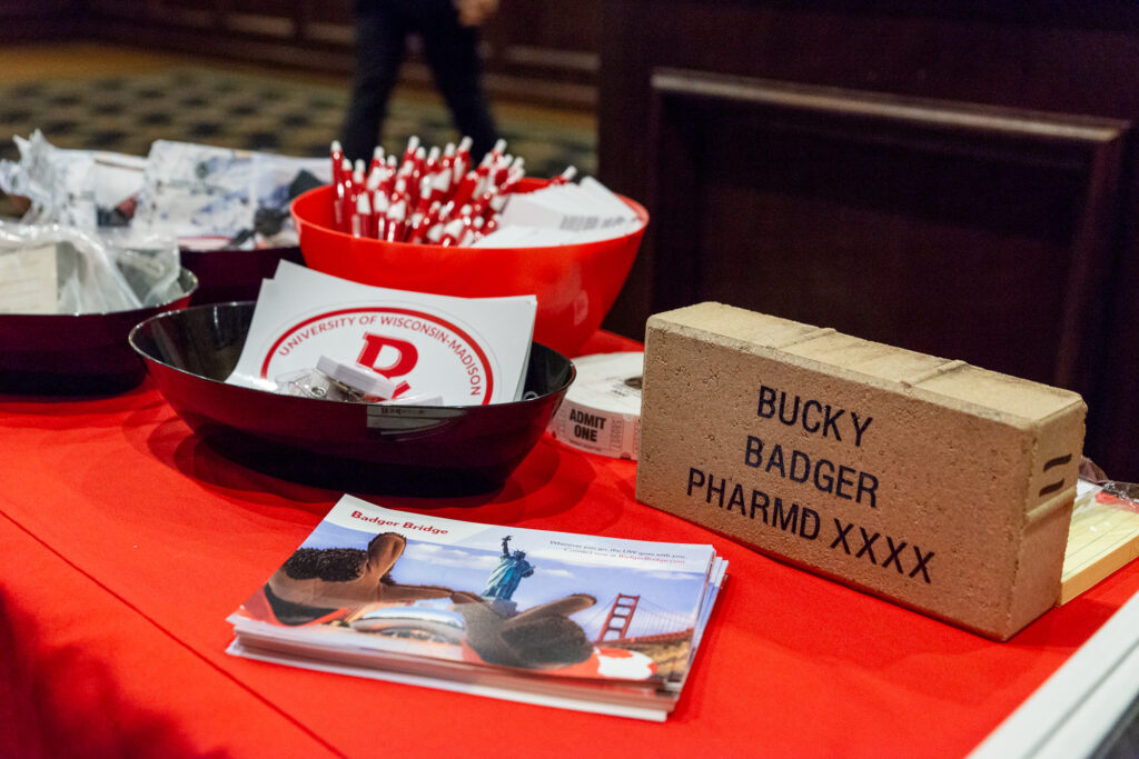 UW-Pharm-Maggianos_20220426__DSC2457_020 Table filled with various handouts like pens, tickets, stickers, information on Badger Bridge, and a brick labeled "Bucky Badger PharmD XXXX"