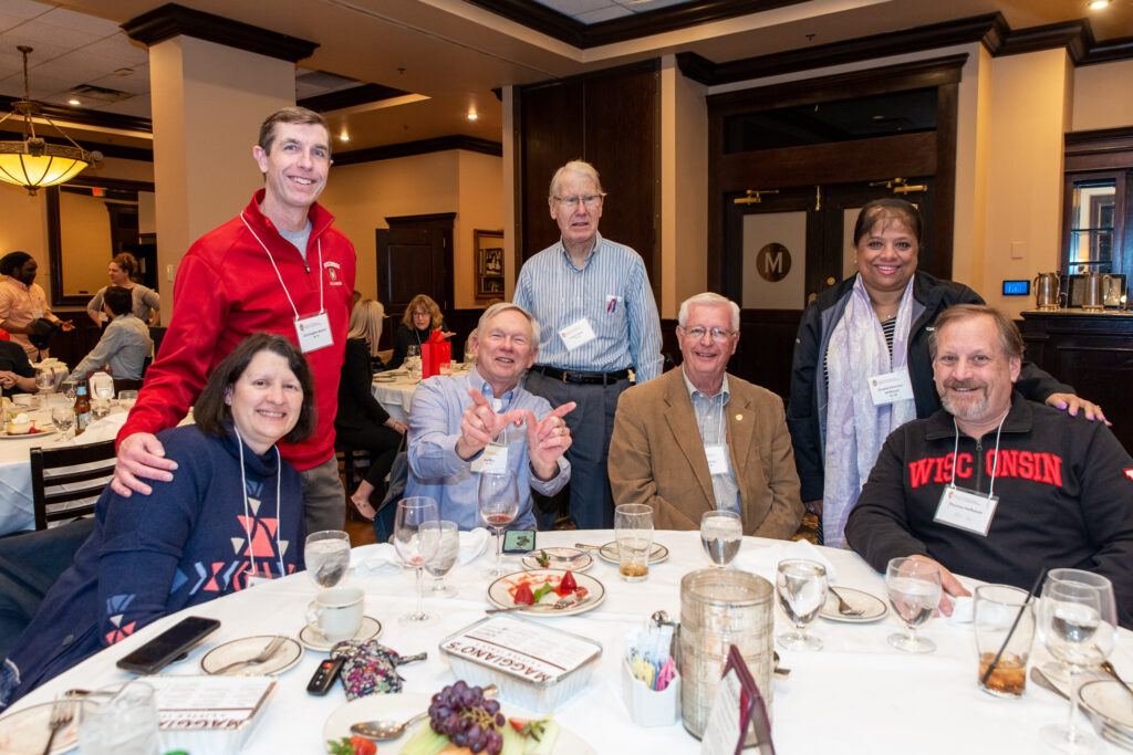 UW-Pharm-Maggianos_20220426__DSC2533_122 Alumni standing and sitting behind a table together, one of the alumni in the middle is forming a 'W' with his hands