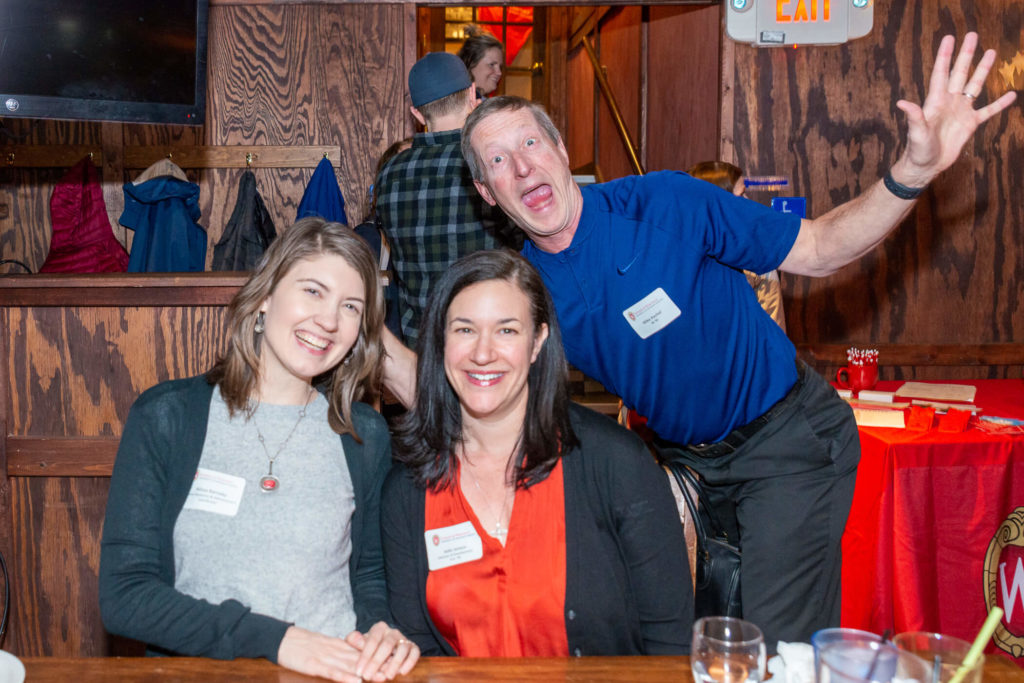 Alissa Karnaky and Julie Jensen sitting at table together with Mike Kachel behind them splaying out his arms and making a funny face