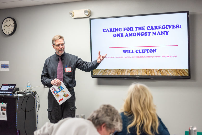 Man speaking and gesturing at front of the room for caregiver event with screen reading "Caring for the Caregiver: one amongst many"