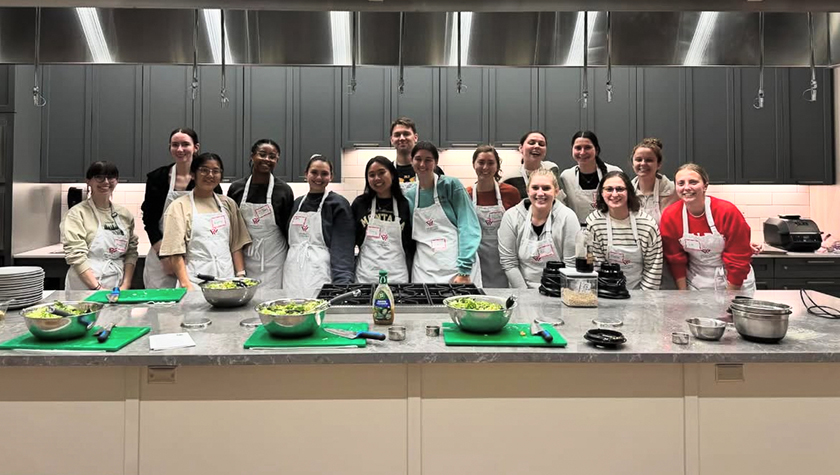 A group of PharmD students in white aprons pose behind a long counter holding several salad bowls and a stovetop.