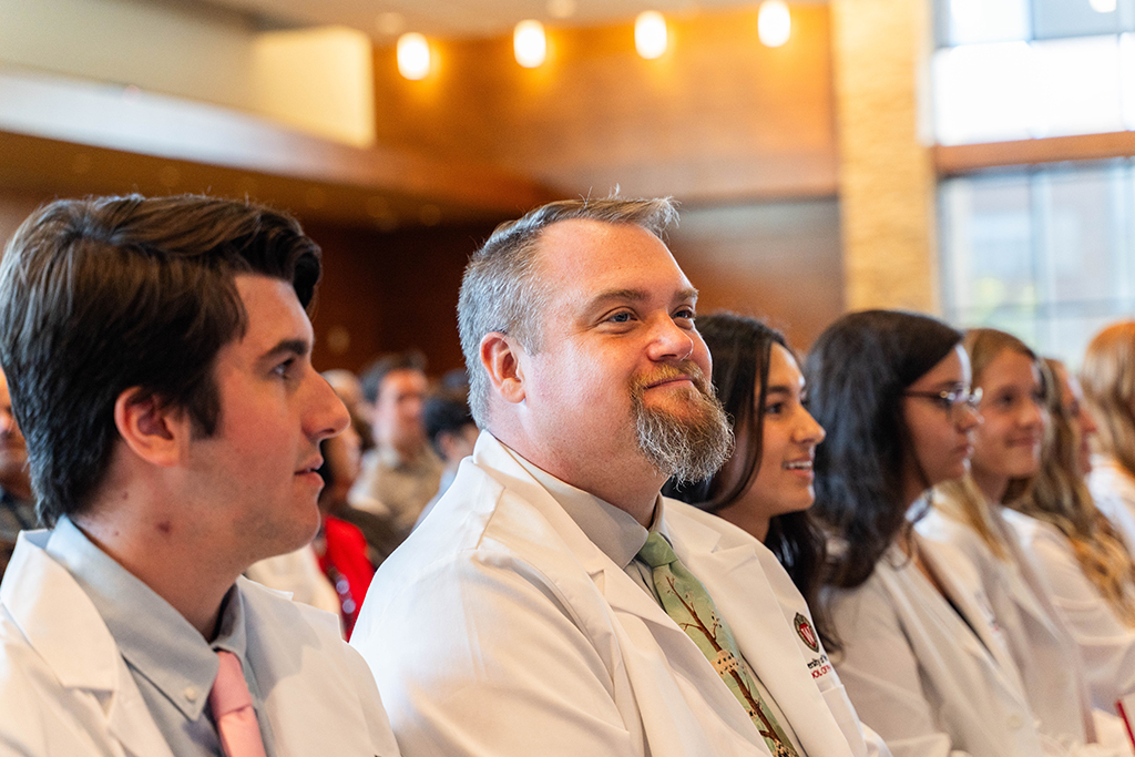 A smiling John Vetterli sits between classmates and listens to speakers during the white coat ceremony.