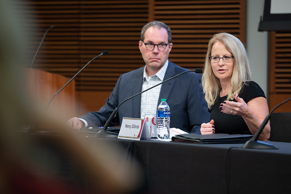 Pat Cory and Nancy Gilbride sit at a table with microphones. Nancy is speaking into his microphone.