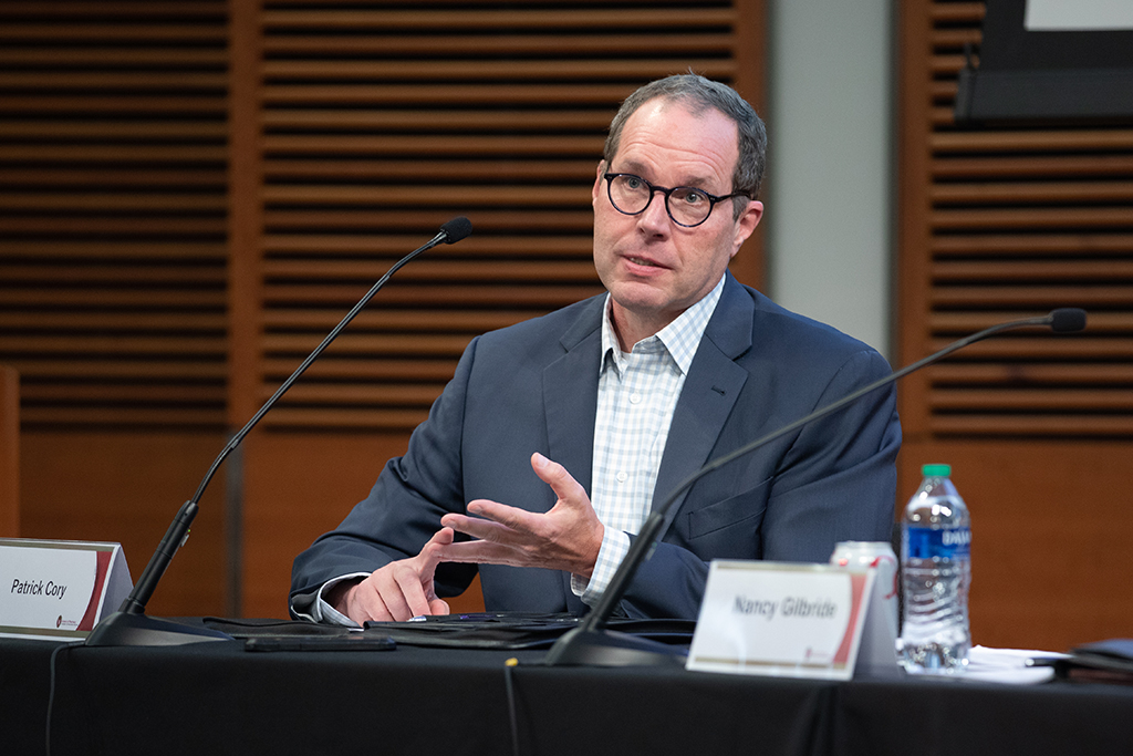 Pat Cory sits at table with a microphone, talking with his hands.