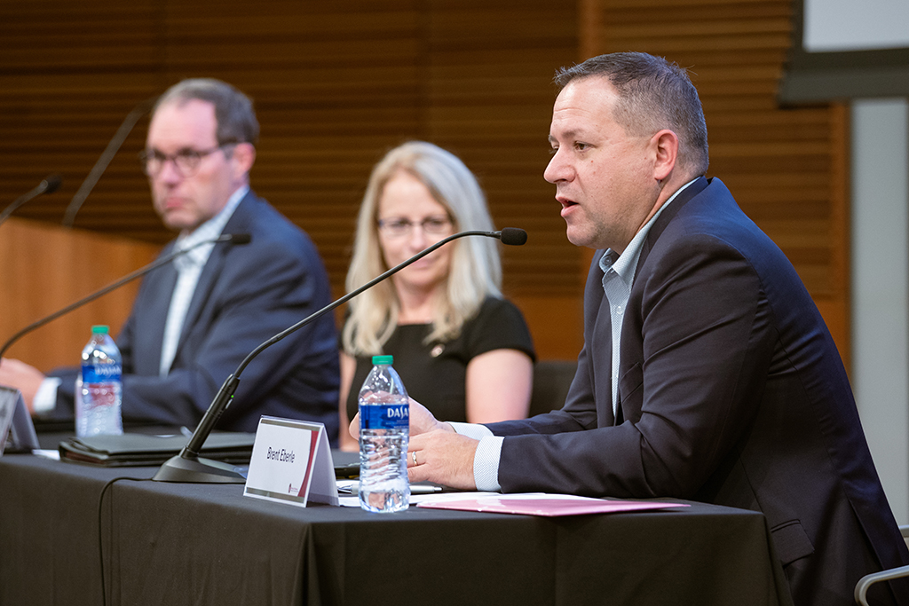 Pat Cory, Nancy Gilbride, and Brent Eberle sit at a table with microphones. Brent is speaking into his microphone.