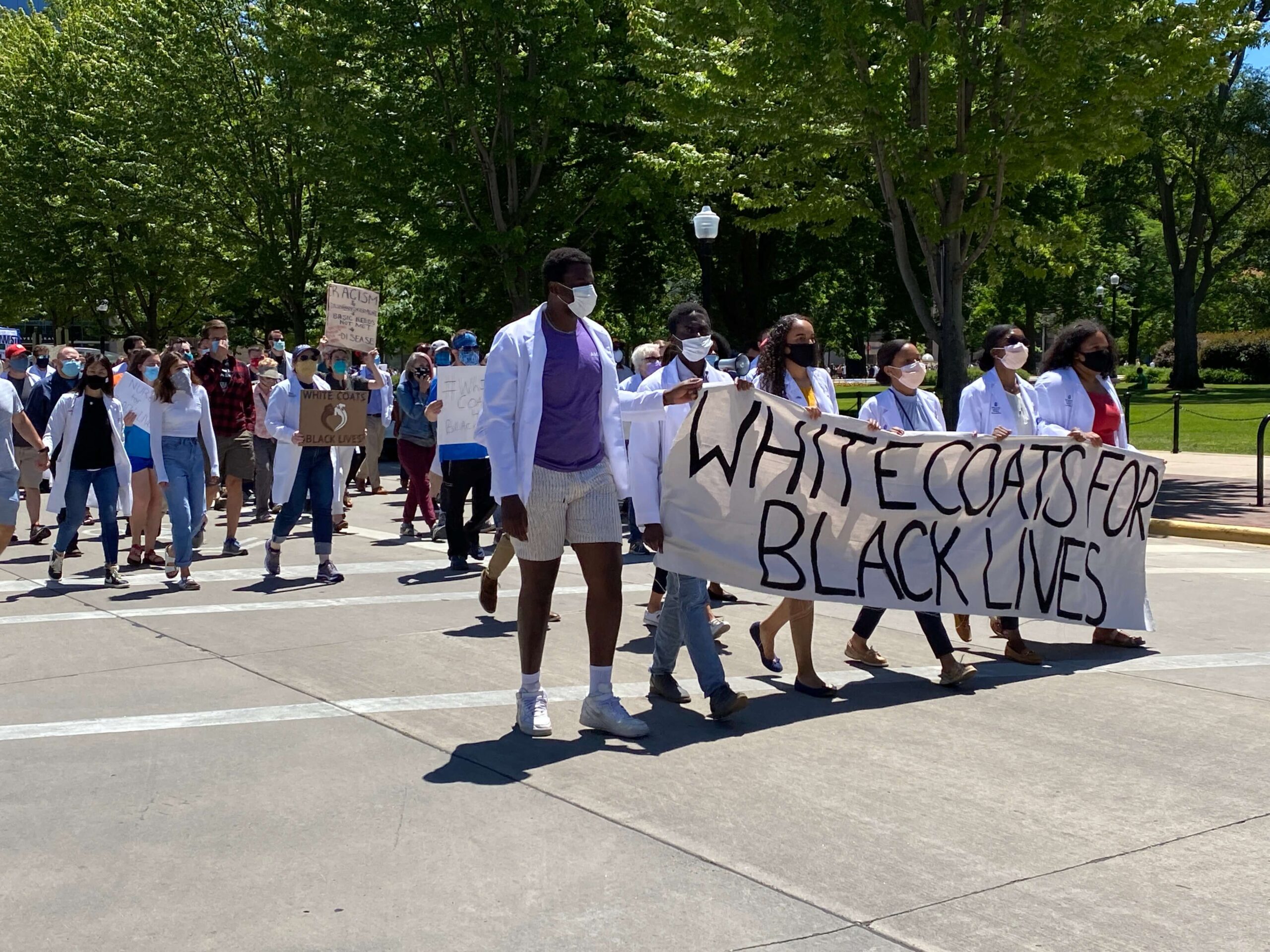 march_IMG_5855 Pharm students wearing white coats and masks walking in protest with a sign reading "White Coats for Black Lives"