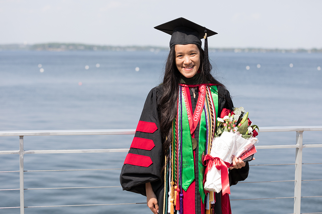 PharmD Class of 2022 graduate Stephaney Cheng poses in her cap and gown with a bouquet of flowers in front of Lake Mendota following the Hooding Ceremony