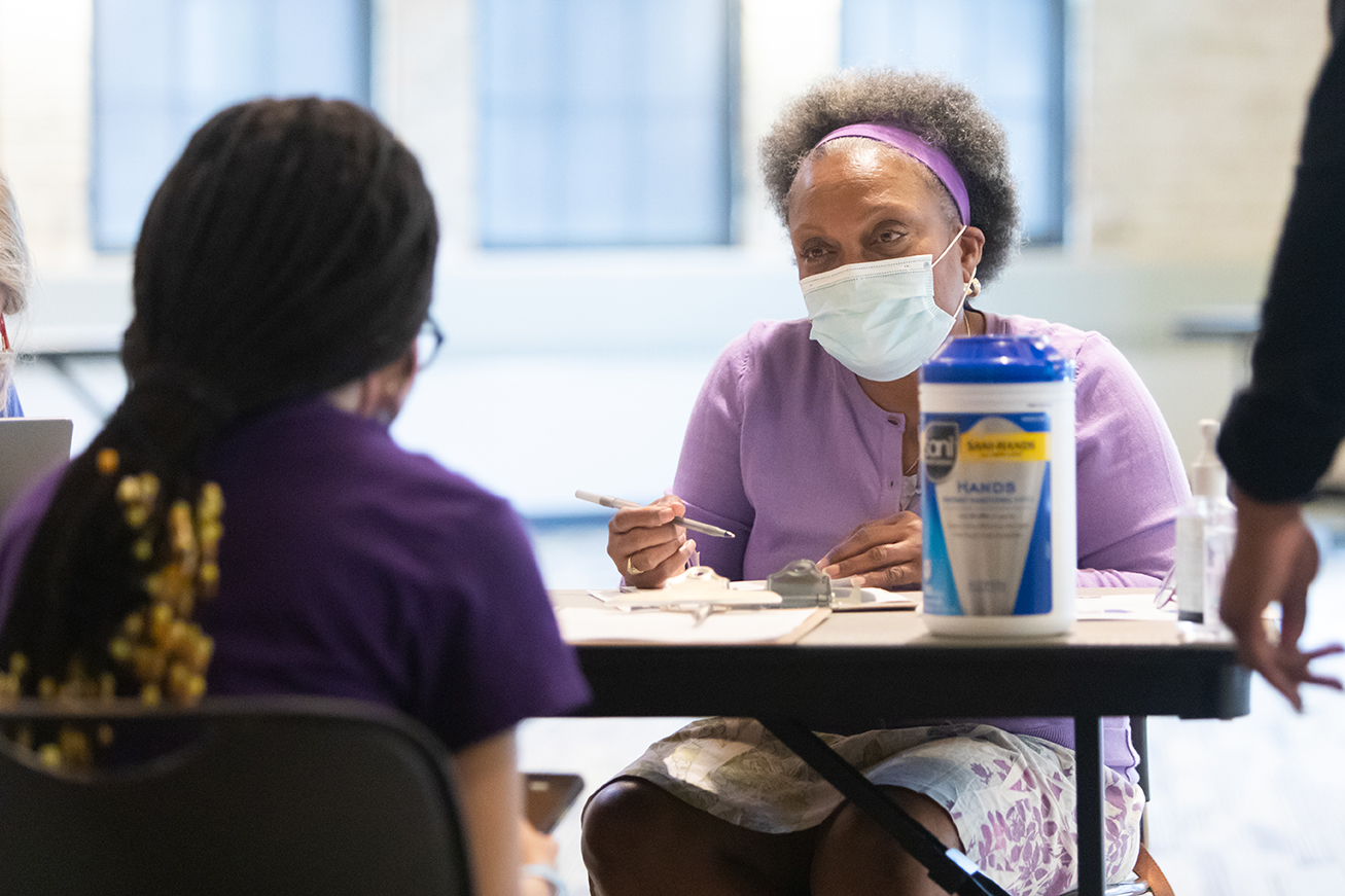 Eva Vivian sitting at table with pen and clipboard in hand, talking to patient across table