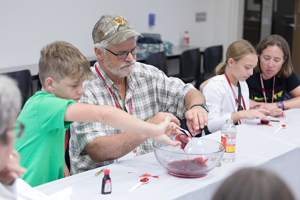 Two attendees pour small cups of red liquid into a bowl