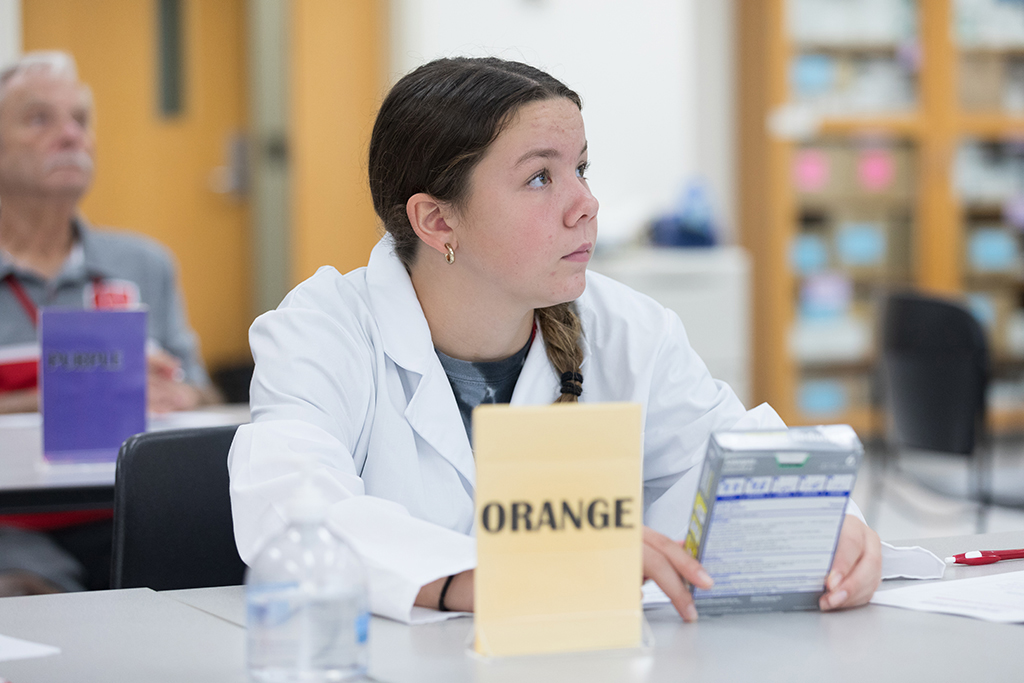 A young attendee holds a box of over-the-counter medication and listens to a speaker