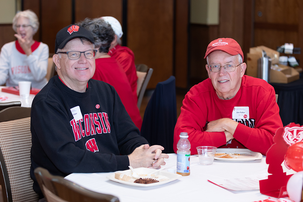 UW–Madison School of Pharmacy 2024 Alumni Tailgate and Watch Party Jim Fidler and another attendee smile while seated at a table, wearing Wisconsin sweatshirts.