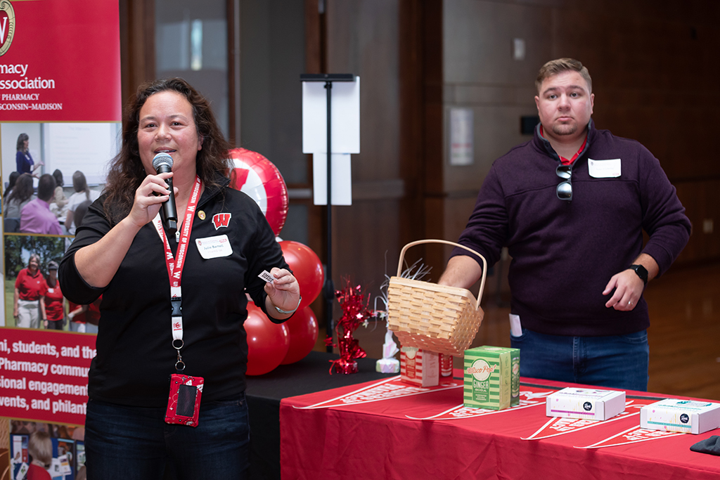 UW–Madison School of Pharmacy 2024 Alumni Tailgate and Watch Party Julie Bartell speaks into a microphone in front of the table of raffle items, Tyler Prickette stands behind the table.