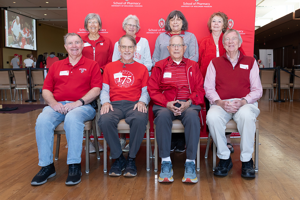 UW–Madison School of Pharmacy 2024 Alumni Tailgate and Watch Party A group of alumni pose for a photo in front of a red School of Pharmacy background, including Russ Jensen, David Duehring, and others.