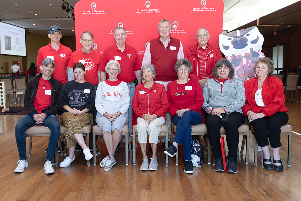 UW–Madison School of Pharmacy 2024 Alumni Tailgate and Watch Party A large group of alumni pose for a photo in front of a red School of Pharmacy background, with a cardboard cutout of Bucky, including Russ Jensen, David Duehring, and others.