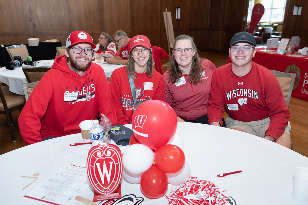 UW–Madison School of Pharmacy 2024 Alumni Tailgate and Watch Party Garrett and Jordan Mattson and Paige and Carl Semrow smile for a photo while sitting at a table decorated with Wisconsin balloons
