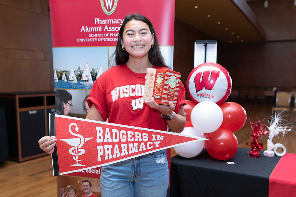 UW–Madison School of Pharmacy 2024 Alumni Tailgate and Watch Party A PharmD students holds up a Badgers in Pharmacy pennant flag and a box of Wisco Pop Strawberry Soda