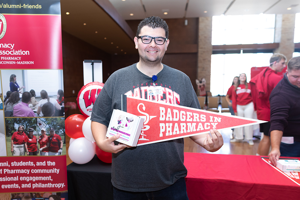 UW–Madison School of Pharmacy 2024 Alumni Tailgate and Watch Party PharmD student Joey Jimenez holds up a Badgers in Pharmacy pennant flag and a box kit for making tassel charms