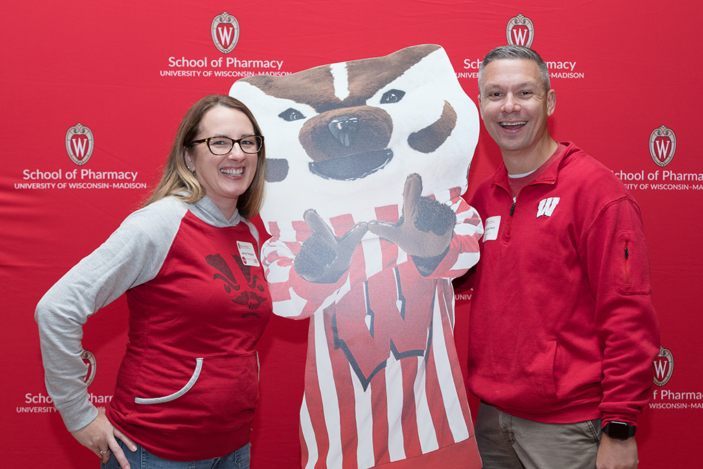 School of Pharmacy Alumni Tailgate Party 2023 Alumni posing next to Bucky Badger cardboard cutout in front of red School of Pharmacy backdrop