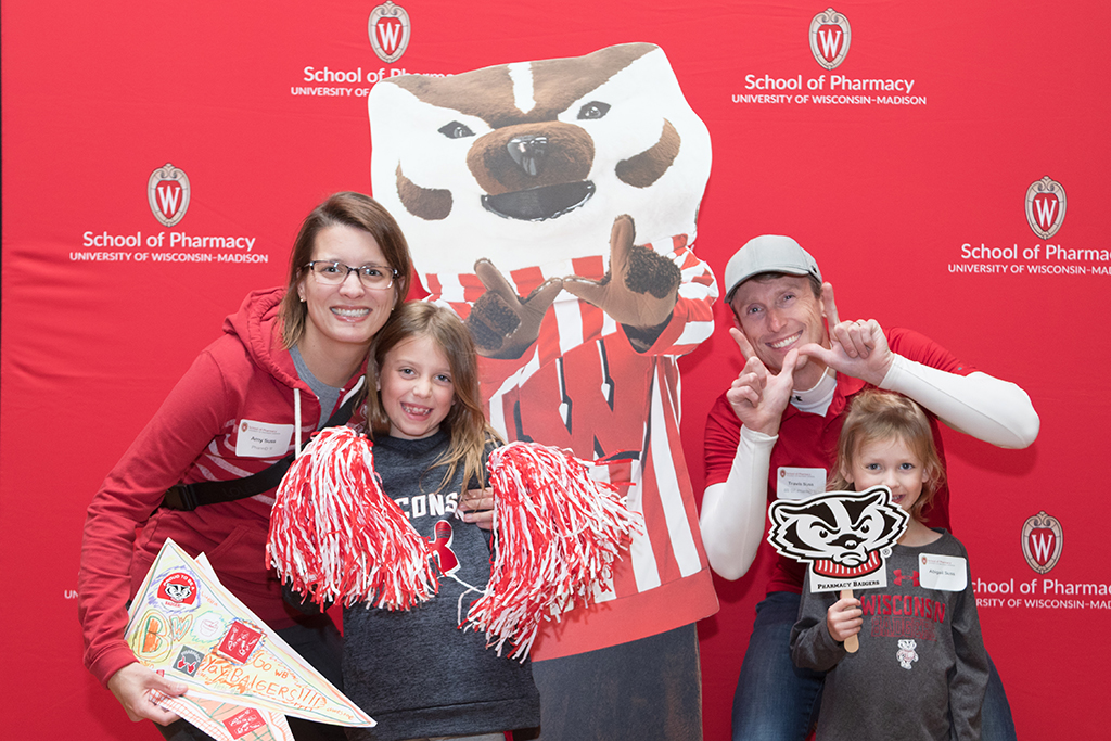 School of Pharmacy Alumni Tailgate Party 2023 Alumnus with spouse and two young daughters posing with Bucky cardboard cutout