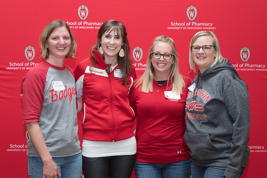 School of Pharmacy Alumni Tailgate Party 2023 Alumni pose arm-in-arm in front of red School of Pharmacy logo background