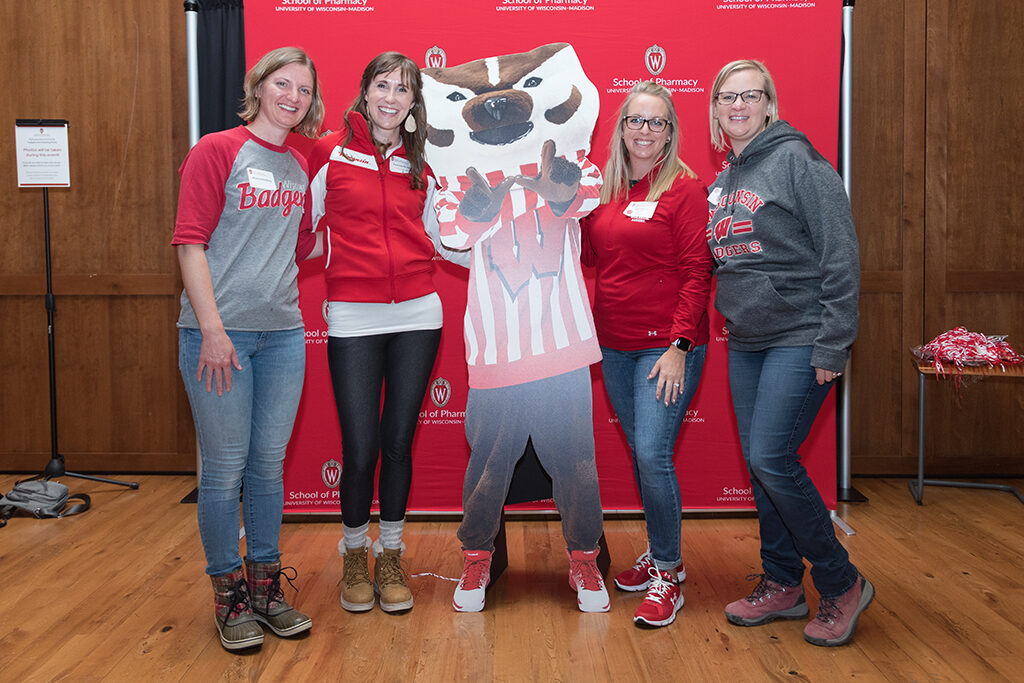 School of Pharmacy Alumni Tailgate Party 2023 Alumni pose with Bucky cardboard cutout in front of red School of Pharmacy logo background