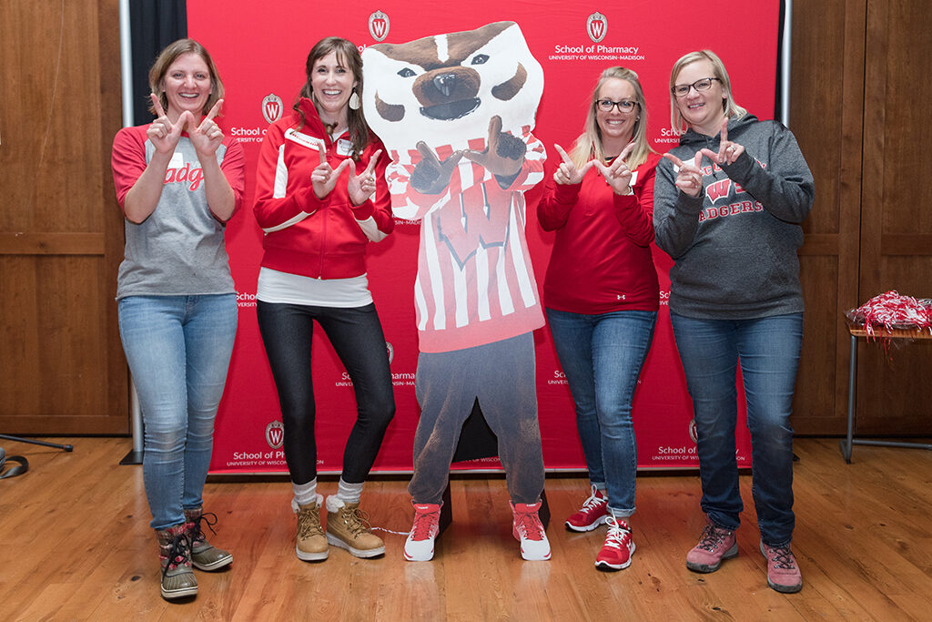 School of Pharmacy Alumni Tailgate Party 2023 Alumni hold up "W"s with Bucky cardboard cutout in front of red School of Pharmacy logo background